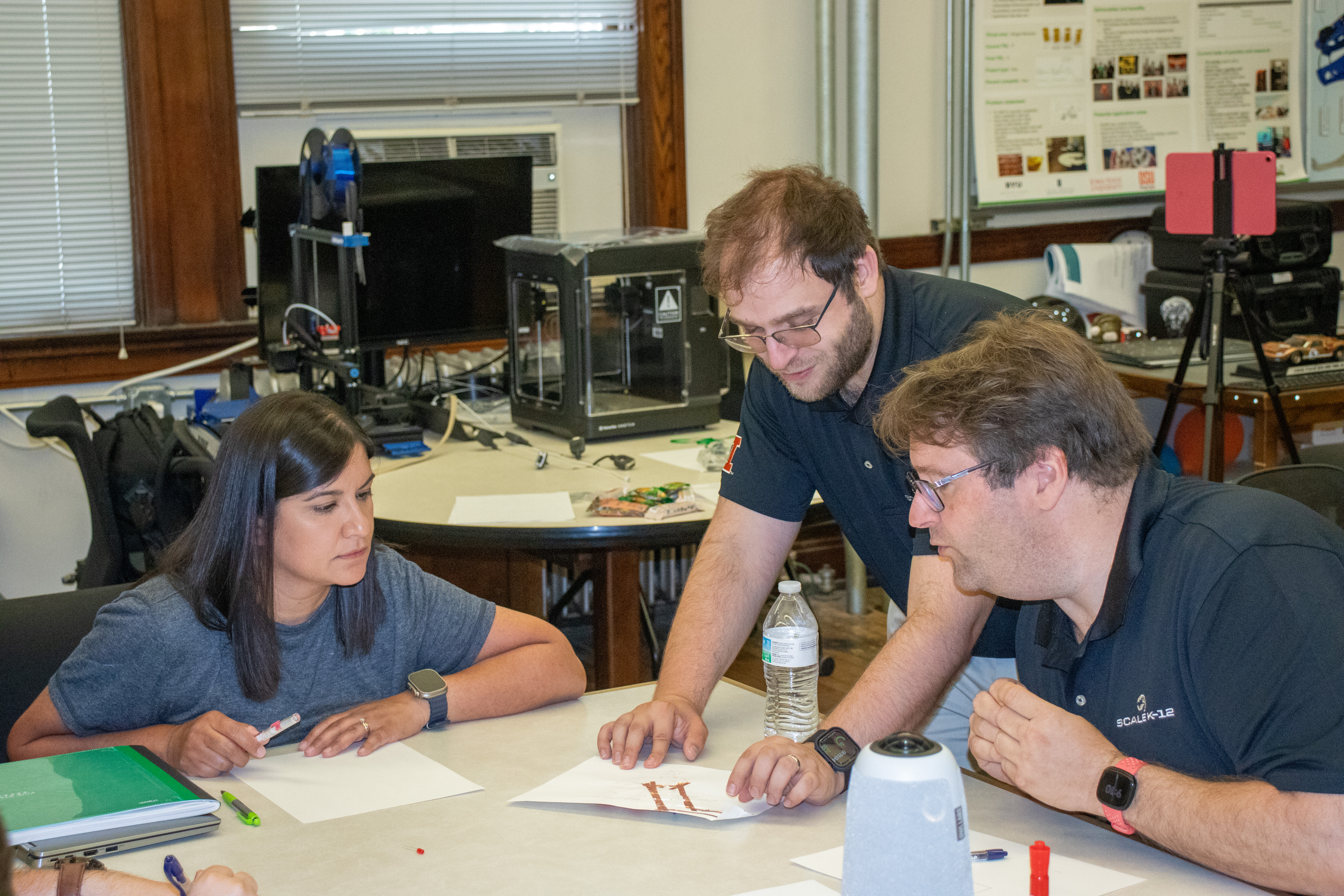 three people. two sitting at a table while one stands at the table demonstrates an activity on a piece of paper