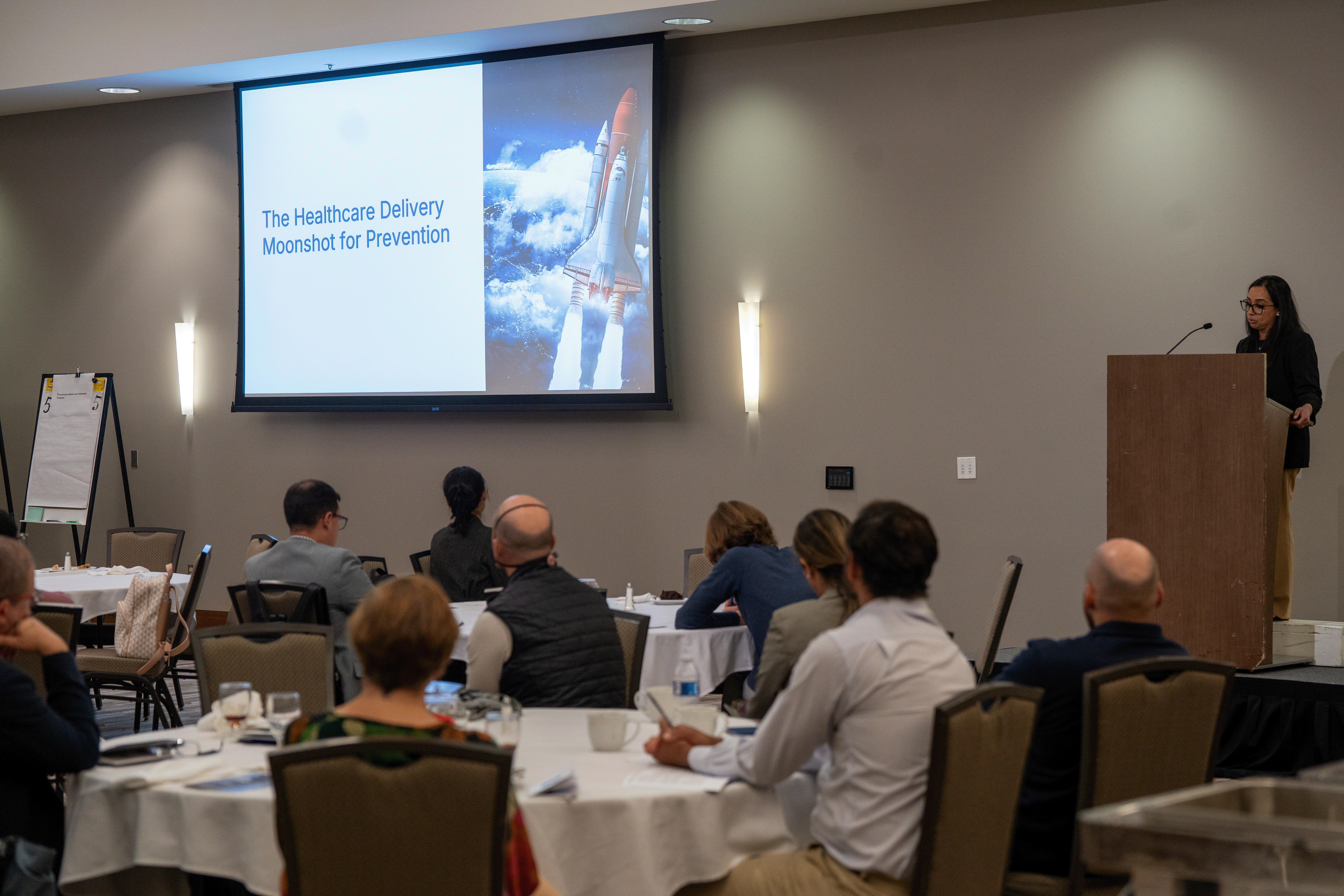 keynote speaker presenting at a podium while people seated at tables listen and look at a powerpoint on a screen